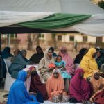 Women and children in colorful attire sit outdoors under a canopy at a community event.