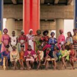 A group of women in vibrant traditional clothing poses under a decorated overpass.